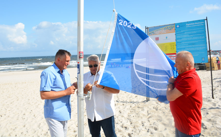 Trzech mężczyzn stoi na plaży obok masztu, przywiązując flagę do liny. W tle morze.