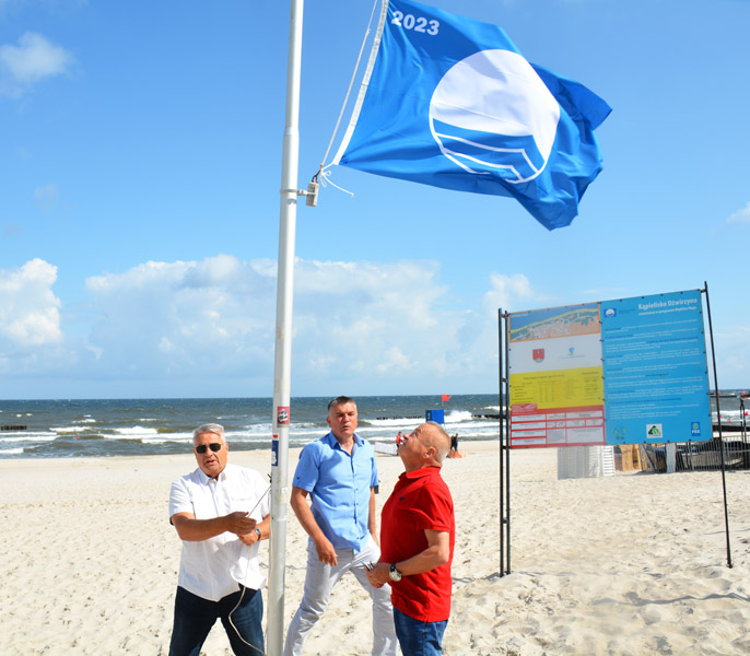 Trzech mężczyzn stoi na plaży, wciągają flagę na maszt. W tle morze.