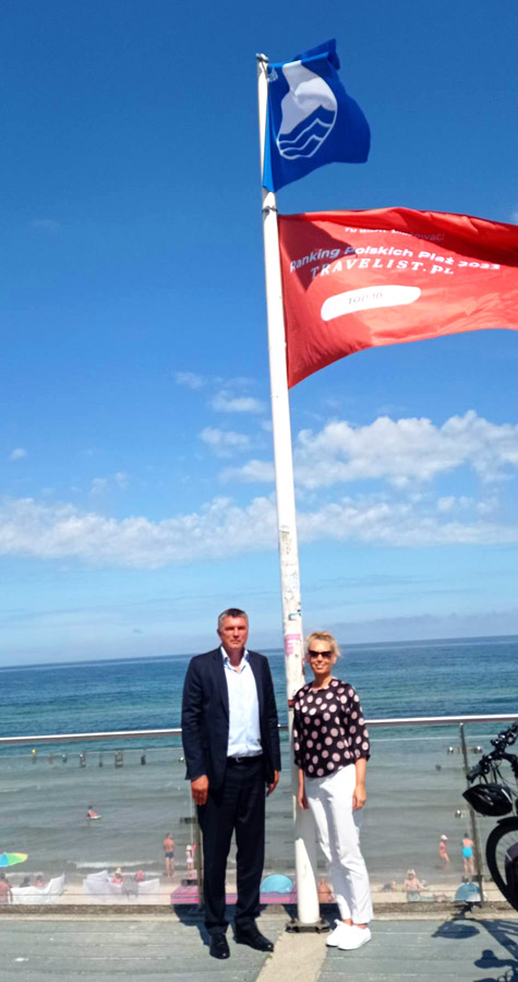 Na punkcie widokowym stoją dwie osoby - kobieta i mężczyzna. Obok nich jest maszt, na którym znajdują się dwie flagi. w tle morze.