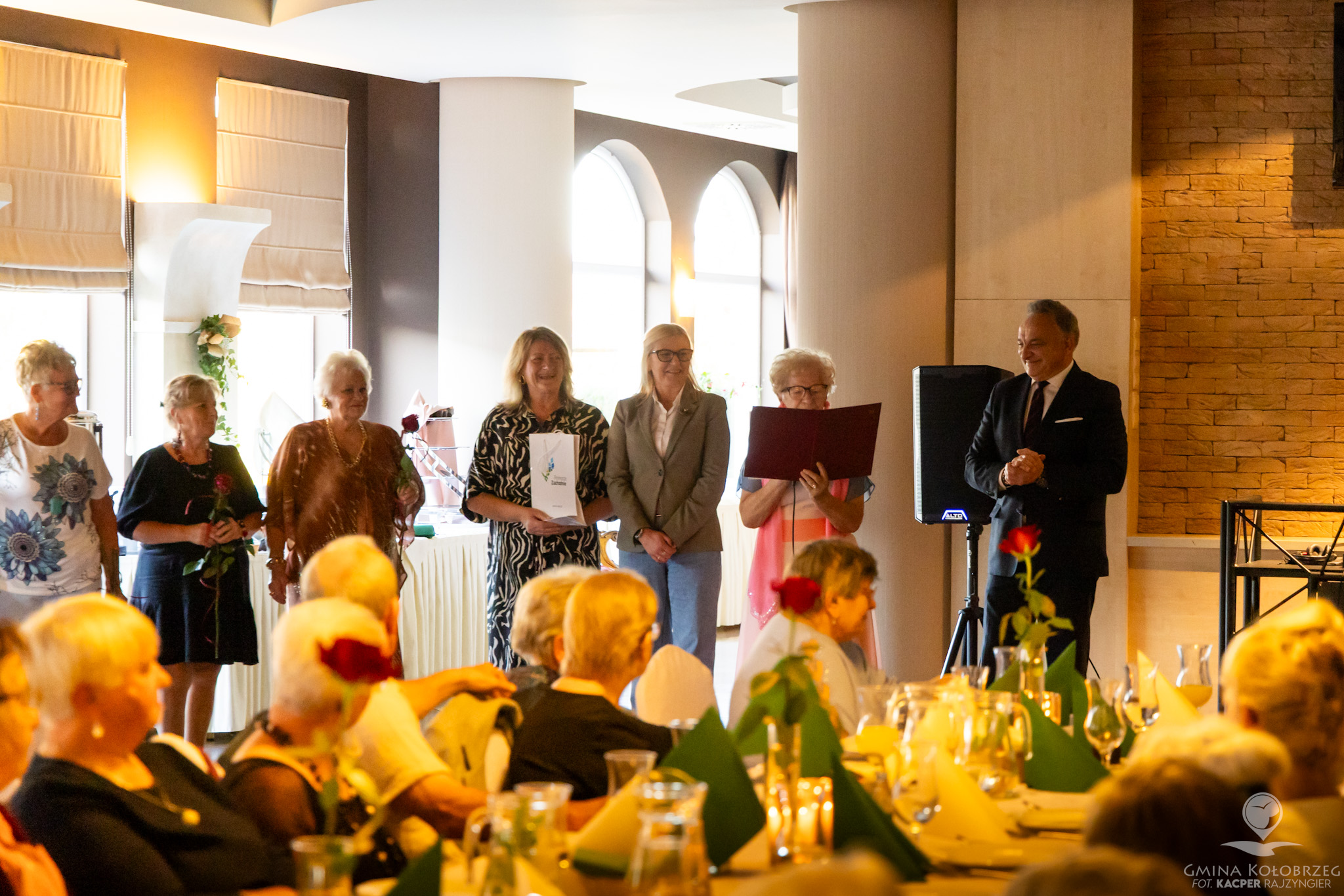 Zdjęcie przedstawia uroczyste rozpoczęcie Balu Seniora. Podczas oficjalnego otwarcia wydarzenia wręczono podziękowania i kwiaty. Elegancka sala, odświętna atmosfera oraz licznie zgromadzeni seniorzy podkreślają wyjątkowy charakter spotkania.
